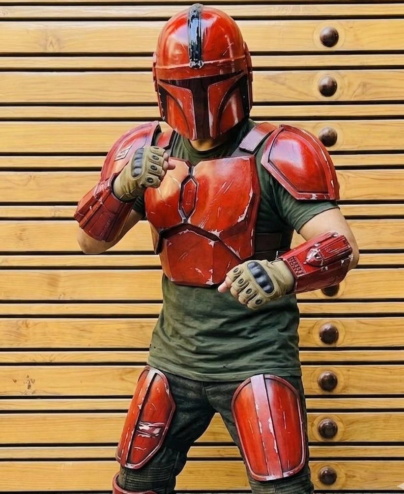 May include: A red Mandalorian armor set, including a helmet, chest plate, shoulder pads, arm guards, and knee pads. The armor has a weathered appearance, suggesting battle. The person is wearing a green shirt and gloves. The background is a wooden wall.