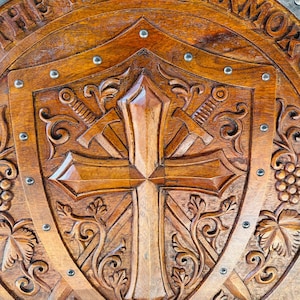 May include: A circular wooden shield featuring a carved cross and two crossed swords in the center. The border includes the text THE WHOLE ARMOR and EPH 6:11. The shield has a metallic outer rim secured with silver-colored metal rivets.