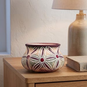 May include: A small, rounded ceramic bowl featuring a decorative pattern of stylised flowers. The exterior displays a gradient of cream, green, and deep maroon tones, with dark maroon centres and outlines defining the floral petals across the surface.
