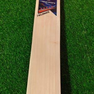 May include: A light-colored wooden cricket bat featuring a white handle grip and a rectangular sticker on the blade. The sticker displays the text YAGAMI SPORTS, FIELDMATE, and ENGLISH WILLOW, along with a logo and five red stars on a dark background.