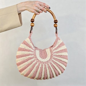 May include: A crescent-shaped crochet handbag featuring alternating vertical stripes of dusty rose and cream yarn. The bag includes a curved wooden handle accented with two dark brown wooden beads on each side, connecting to the main crocheted body.