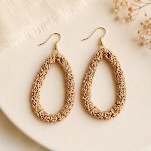 Peut inclure: Une paire de boucles d'oreilles ovales ouvertes avec une texture tissée de couleur beige clair. Les boucles d'oreilles ont des crochets dorés et sont présentées sur une surface blanche. Les boucles d'oreilles sont de style bohème.