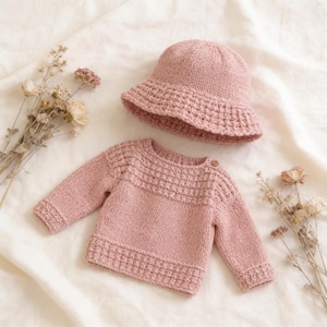 Könnte beinhalten: Ein altrosa gestrickter Langarmpullover und ein passender Fischerhut. Der Pullover hat ein strukturiertes Waffelstrickmuster an der Passe, den Bündchen und am Saum sowie einen kleinen Holzknopf am Ausschnitt. Der Hut hat eine breite Krempe und Textur.
