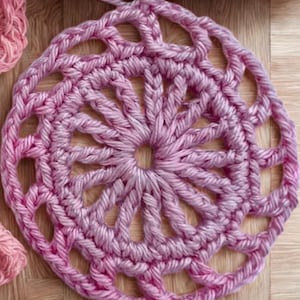 Op de afbeelding: Close-up van een gehaakte doily in roze tinten. Het cirkelvormige ontwerp heeft een openwerkpatroon met een centrale ster en stralende spaken. Een haaknaald is in het werkstuk gestoken, met een stuk garen zichtbaar. De doily ligt op een houten oppervlak.