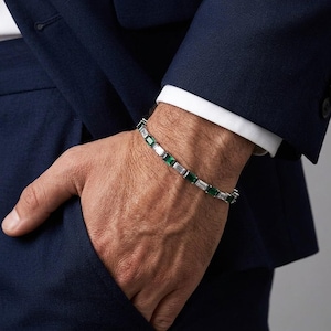 Könnte beinhalten: Ein silbernes Armband mit Smaragden und weißer Baguette-Schliff-Steinen. Das Armband wird am Handgelenk getragen, mit einem marineblauen Anzug im Hintergrund. Das Armband ist ein Schmuckstück.