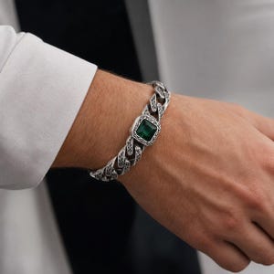 May include: A silver-toned curb chain bracelet featuring a central square-cut emerald green gemstone. The metal links are decorated with intricate, engraved scrollwork patterns throughout the entire length of the chain, creating a textured appearance.