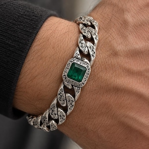 May include: A silver curb chain bracelet featuring a central rectangular emerald green gemstone set in a decorative frame. Each individual link of the chain is engraved with intricate floral patterns, creating a textured and detailed appearance.