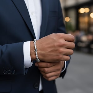 May include: A silver bracelet with alternating clear stones and black clover-shaped accents. The bracelet is worn on a wrist, with a navy blue suit and white dress shirt in the background.