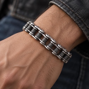 May include: A silver-toned metal bracelet designed to resemble a motorcycle drive chain. The links feature a dark, oxidized finish with intricate, etched patterns on the central rollers, creating a textured and industrial appearance on the wrist.
