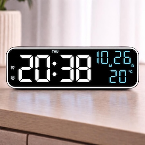May include: A black digital alarm clock displaying the time as 20:38, with the date 10.26 and the temperature 20°F. The clock has a rectangular shape with rounded corners and a white border. The display is lit with blue and white digits.