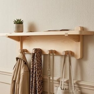 Peut inclure: Une étagère murale en bois clair avec un porte-manteau intégré. L'étagère contient une petite plante en pot et quelques petits objets. En dessous, trois chevilles en bois maintiennent un manteau beige, une écharpe à motifs, une laisse et un sac fourre-tout.