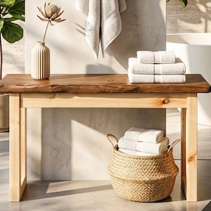 Puede incluir: Un banco de madera con una parte superior de borde vivo y patas de color claro. El banco contiene toallas blancas dobladas, un jarrón decorativo con una flor seca y una cesta tejida llena de toallas. Un ambiente de baño de tonos neutros.