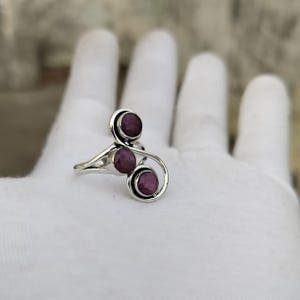 Puede incluir: Un anillo de plata con tres piedras redondas facetadas de color rojo rosado intenso, engastadas en una banda de metal calada y en espiral. El diseño presenta un estilo bypass donde el metal se curva alrededor de cada piedra individualmente.