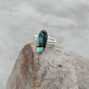 Puede incluir: Un anillo de plata con una incrustación ovalada de concha de abulón en tonos iridiscentes verdes, azules y negros. La banda del anillo está elaborada con un diseño de hilo de plata trenzado, proporcionando una apariencia texturizada.