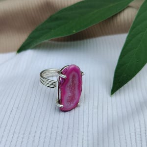 May include: A silver ring featuring a vertical, oval-shaped hot pink druzy agate stone. The stone has a textured, crystalline center and is held in place by four silver prongs. The ring band consists of three thin, parallel silver wires.