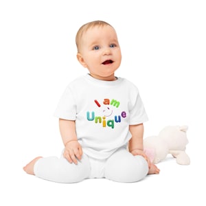 Könnte beinhalten: Ein Baby in einem weißen T-Shirt und einer Hose. Auf dem T-Shirt steht in bunten Buchstaben "I am Unique". Ein Stofftier liegt neben dem Baby.