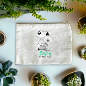 Könnte beinhalten: Cremefarbene Canvas-Reißverschlusstasche mit einem niedlichen Cartoon-Frosch, der einen Löwenzahn hält. Der Text "Enjoy the little things" ist darunter in Türkis und Schwarz gedruckt.