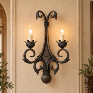 Puede incluir: Un aplique de pared de metal color bronce oscuro con dos luces tipo vela y cubiertas color crema. El accesorio presenta un eje central vertical con volutas decorativas, bases para velas en forma de hoja y una base esférica con textura.