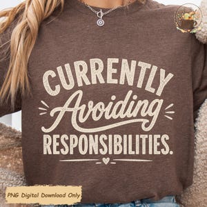 May include: A brown crewneck sweatshirt featuring the cream-colored text "CURRENTLY Avoiding RESPONSIBILITIES." The design includes decorative lines and a small heart icon centered below the text. The fabric has a heathered texture throughout.