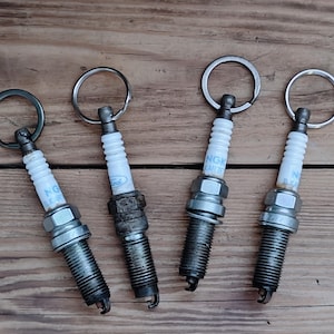 Könnte beinhalten: Vier Zündkerzen-Schlüsselanhänger aus Metall, die in einer Reihe angeordnet sind. Jede Zündkerze verfügt über einen weißen Keramikisolator mit blauem Text, einen metallischen Gewindesockel und einen silberfarbenen Ring zur Befestigung.