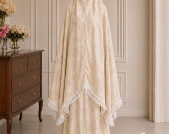 Könnte beinhalten: Beiges Gebetskleidungsset mit floralem Muster. Das Set beinhaltet eine Kopfbedeckung und ein langes Kleid. Weiße Rüschenbesätze akzentuieren die Kanten des kapuzenartigen Oberteils. Der Stoff hat einen dezenten Glanz. Die Frau trägt eine passende Kopfbedeckung.