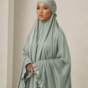 May include: A sage green prayer garment set. The set includes a head covering with a floral embellishment and a matching flowing dress with ruffled sleeves. The fabric drapes softly, creating a modest and elegant silhouette.