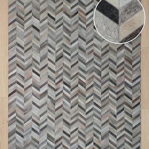 May include: A rectangular area rug featuring a repeating chevron pattern composed of individual hide patches in shades of gray, white, black, and tan. The surface displays a textured, multi-tonal appearance with a consistent geometric arrangement.