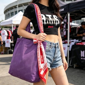 Peut inclure: Un sac fourre-tout violet avec une sangle &agrave; motifs rouges et blancs et un foulard assorti nou&eacute; &agrave; la poign&eacute;e. Le sac est port&eacute; &agrave; l'&eacute;paule. Un crop top noir avec le mot "BTS" et une silhouette de personnes est &eacute;galement visible.