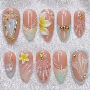 May include: A set of ten artificial nails with a light pink base, each adorned with unique 3D designs. The designs include seashells, starfish, and floral accents in shades of white, yellow, and gold. Some nails feature gold embellishments and pastel color gradients.