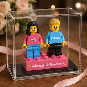 May include: A Lego-style couple figurine set inside a clear display case. The figures, Mia and Marcus, are holding hands and standing on a pink base with the words "Always & Forever". The figures are wearing pink and blue tops and blue pants.