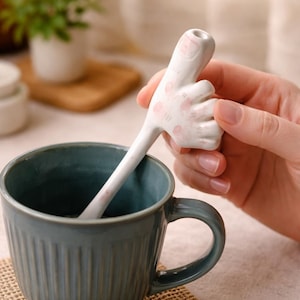 May include: A white ceramic spoon with a hand-shaped handle, featuring pink accents, is placed in a teal mug. The spoon's design includes a thumb and fingers. The mug has vertical ridges and a curved handle. The background is blurred.