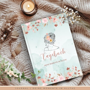 Puede incluir: Un diario con una cubierta azul claro con un diseño floral y una ilustración de la silueta de una mujer. La palabra "Tagebuch" está escrita en cursiva. El diario está rodeado por una vela y flores secas.