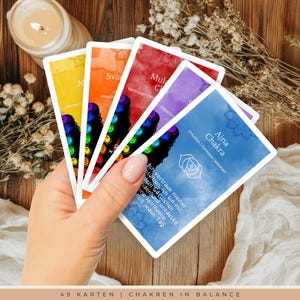 Könnte beinhalten: Eine Hand hält einen Fächer aus bunten Chakra-Karten. Jede Karte hat eine andere Farbe und ein anderes Design mit Text auf Deutsch. Die Karten sind mit Chakra-Namen und zugehörigen Konzepten beschriftet. Der untere Teil des Bildes lautet "49 KARTEN | CHAKREN IN BALANCE."