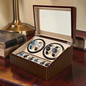 Op de afbeelding: Een bruine houten horlogedoos met een glazen deksel, met daarin diverse polshorloges. De doos heeft een lichtgekleurd interieur en ronde horlogehouders. Sommige horloges hebben leren bandjes en zilveren of blauwe wijzerplaten.