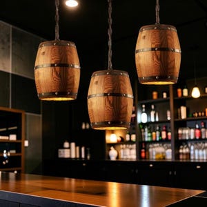 May include: Three wooden barrel pendant lights suspended from chains. The lights emit a warm glow, with metal bands encircling the barrels. The background reveals a bar setting with shelves of bottles and a wooden countertop.
