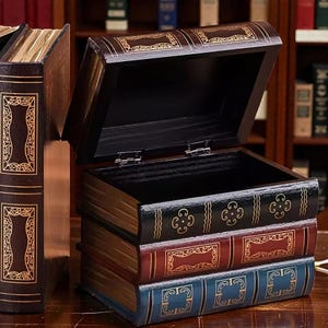 Può includere: Una scatola di legno progettata per assomigliare a una pila di libri, con il coperchio aperto. La scatola è dipinta nei toni del marrone, nero, rosso e blu, con accenti dorati. L'interno della scatola è nero.