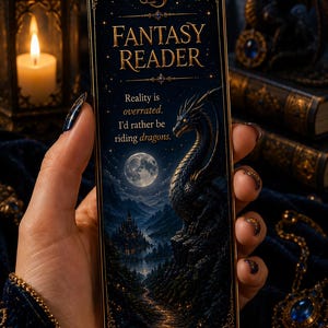 Könnte beinhalten: Ein rechteckiges Lesezeichen mit dunkelblauem und goldenem Design, das eine Drachenillustration, einen Vollmond und eine Schlosslandschaft zeigt. Der Text lautet FANTASY READER und Reality is overrated. I'd rather be riding dragons. Es hat goldene Filigranränder.