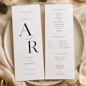 Könnte beinhalten: Zwei weiße Hochzeitskarten mit schwarzer Serifenschrift und kursiver Typografie. Die linke Karte zeigt die Initialen A und R, die Namen Alyssa und Robert, das Datum 06-06-2026 und New York. Die rechte Karte listet den Ablauf der Zeremonie auf.