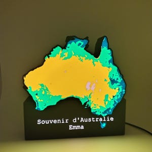 May include: A glowing, illuminated map of Australia featuring a vibrant yellow center surrounded by green and blue coastal regions. The map is mounted on a black rectangular base with the white text Souvenir d'Australie and the name Emma printed on it.
