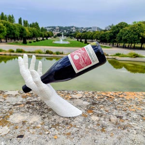 Peut inclure: Une sculpture blanche en forme de main texturée tenant une bouteille de vin en verre foncé. La bouteille présente une étiquette avec un dessin graphique en lignes rouges et le texte Les Grandes Collines. La sculpture a une finition craquelée.
