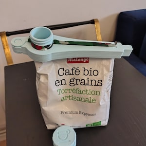Pode incluir: Um clipe de plástico azul-claro para sacos com um bico circular e uma tampa de rosca a condizer, fixado num saco branco de café Malongo Café bio en grains. O clipe apresenta uma tira de tecido com um padrão verde, vermelho e preto.