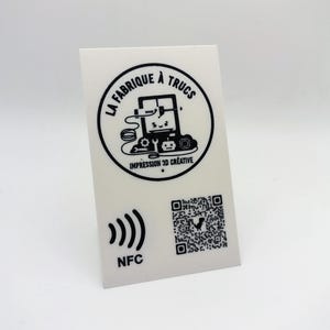 May include: A white rectangular card featuring a black circular logo with a 3D printer graphic and the text LA FABRIQUE À TRUCS IMPRESSION 3D CRÉATIVE. Below the logo is a black NFC signal icon with the text NFC and a square QR code.