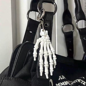 Peut inclure: Un porte-clés en forme de main de squelette en plastique blanc, fixé à un mousqueton et un anneau en métal argenté. La breloque représente une main humaine avec des phalanges articulées et est suspendue à la bandoulière d'un sac noir texturé.