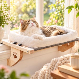 May include: A fluffy white cat bed mounted on a light-colored wooden window perch. The perch features a cutout paw print design on the side supports and black adjustment knobs. The bed has a soft, plush white cushion for resting.