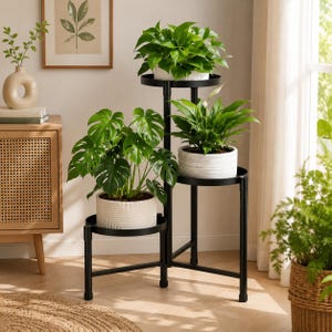 May include: A black metal tiered plant stand with three circular shelves at varying heights. The stand holds three potted plants: a large monstera in a white textured pot, a peace lily in a white ribbed pot, and a leafy green plant on top.