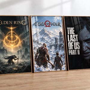 Puede incluir: Tres pósteres verticales enmarcados con arte de videojuegos. El póster de la izquierda muestra Elden Ring con un símbolo dorado brillante. El central muestra God of War con dos figuras en un paisaje nevado. El de la derecha presenta The Last of Us Part II.