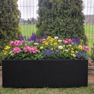 Puede incluir: Una jardinera rectangular negra llena de una variedad de flores coloridas, incluyendo flores rosas, amarillas, blancas y moradas. La jardinera está colocada sobre una zona de césped, con una valla de alambre y árboles en el fondo.