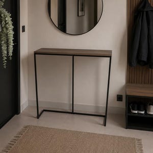 Op de afbeelding: Een moderne entree met een rechthoekige console met een donkerhouten blad en een zwart metalen frame. Een ronde zwarte spiegel hangt boven de tafel. Een geweven vloerkleed en een schoenenrek maken de inrichting compleet.