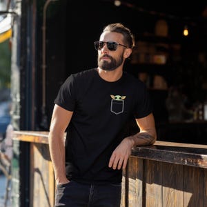 K&ouml;nnte beinhalten: Schwarzes T-Shirt mit wei&szlig;em Taschen-Design mit einer gr&uuml;nen und beigefarbenen Cartoon-Figur. Das T-Shirt ist kurz&auml;rmlig und scheint aus einem weichen Material zu sein. Das Taschen-Design ist auf der Brust zentriert.