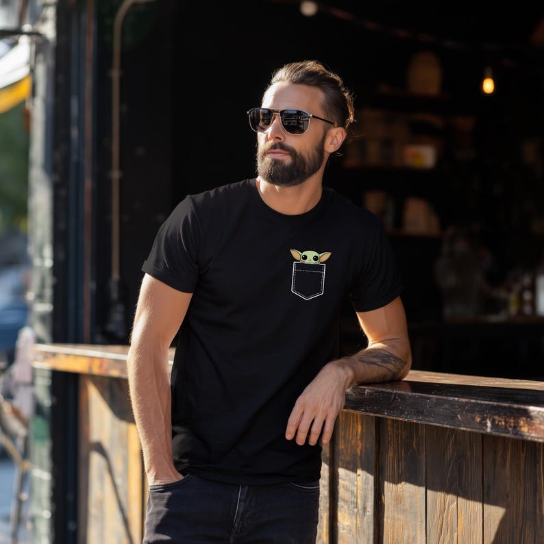 K&ouml;nnte beinhalten: Schwarzes T-Shirt mit wei&szlig;em Taschen-Design mit einer gr&uuml;nen und beigefarbenen Cartoon-Figur. Das T-Shirt ist kurz&auml;rmlig und scheint aus einem weichen Material zu sein. Das Taschen-Design ist auf der Brust zentriert.